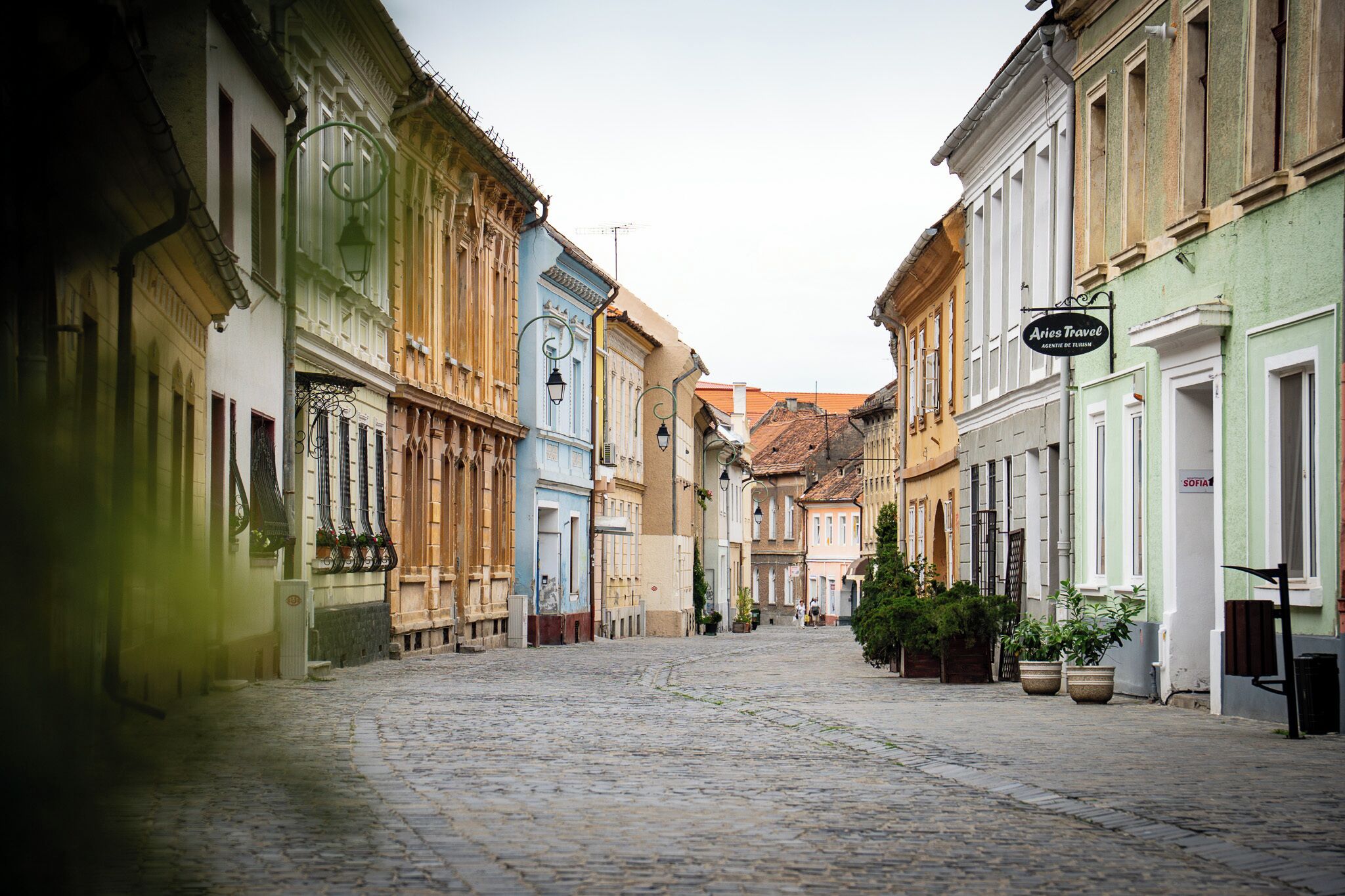 A cobblestone street lined with historic buildings, a sign for 'Ariès Travel', and a few potted plants.