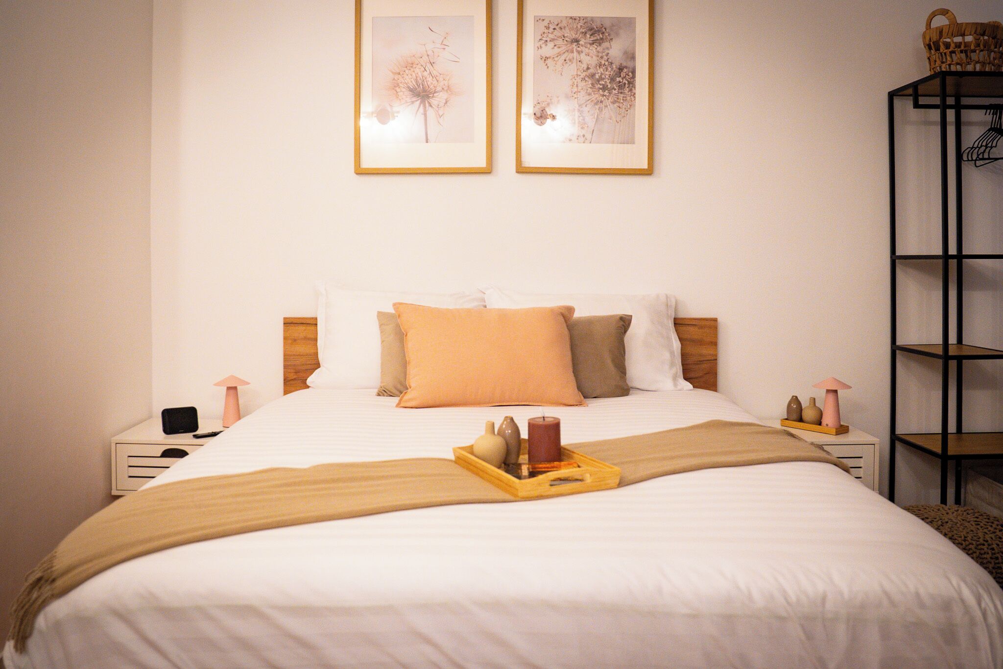 A neatly made bed with a tray of items, two framed pictures on the wall, and a shelf with a basket on the right.
