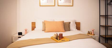 A neatly made bed with a tray of items, two framed pictures on the wall, and a shelf with a basket on the right.