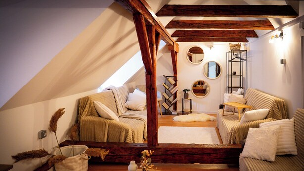 Um quarto de sótão acolhedor, com teto inclinado, estrutura de vigas de madeira, um sofá, uma pequena mesa e um espelho.