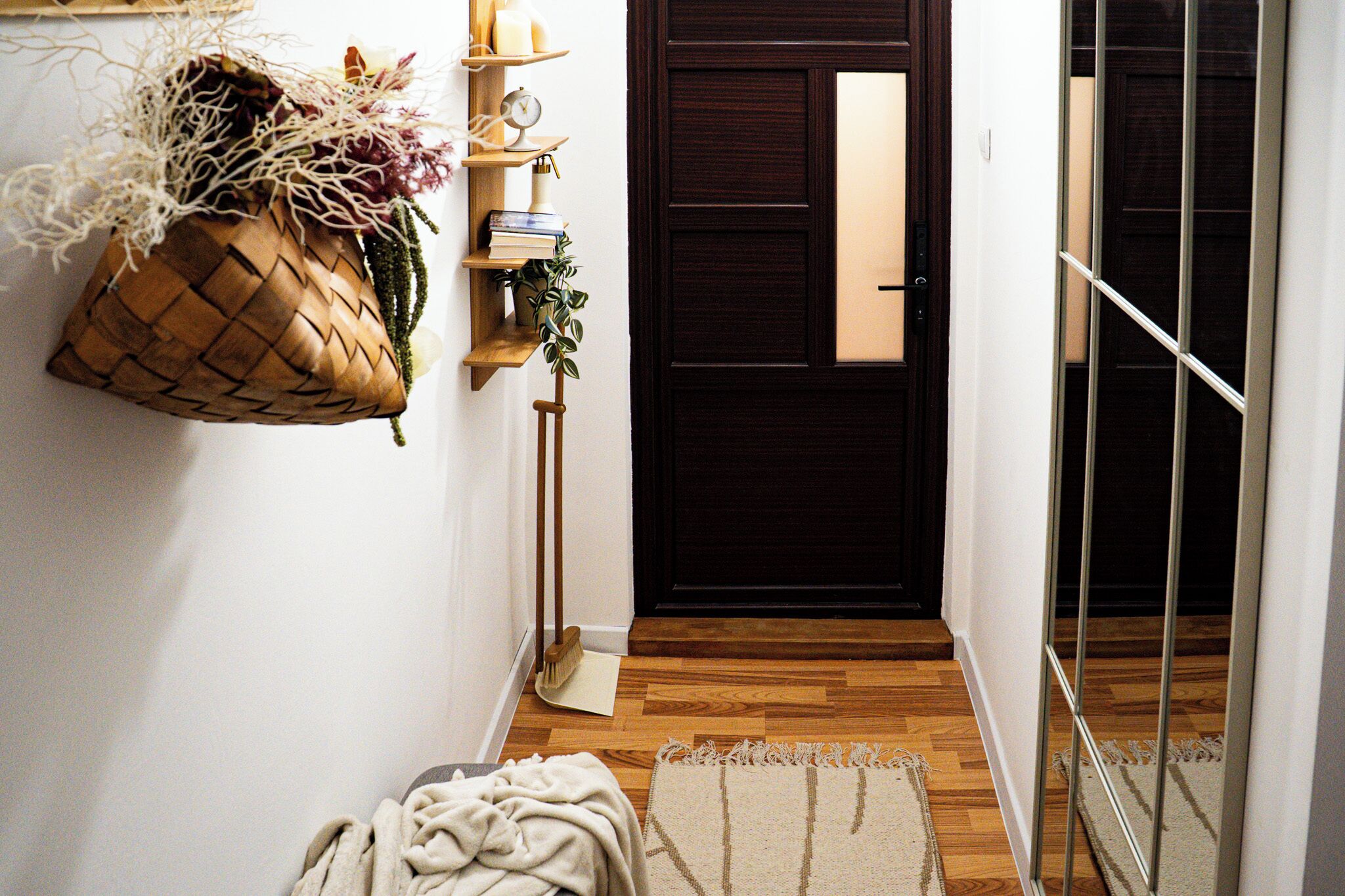 Un pasillo con suelo de madera, una puerta oscura, una cesta colgante con plantas, un estante pequeño con libros y un reloj, y una pared blanca.