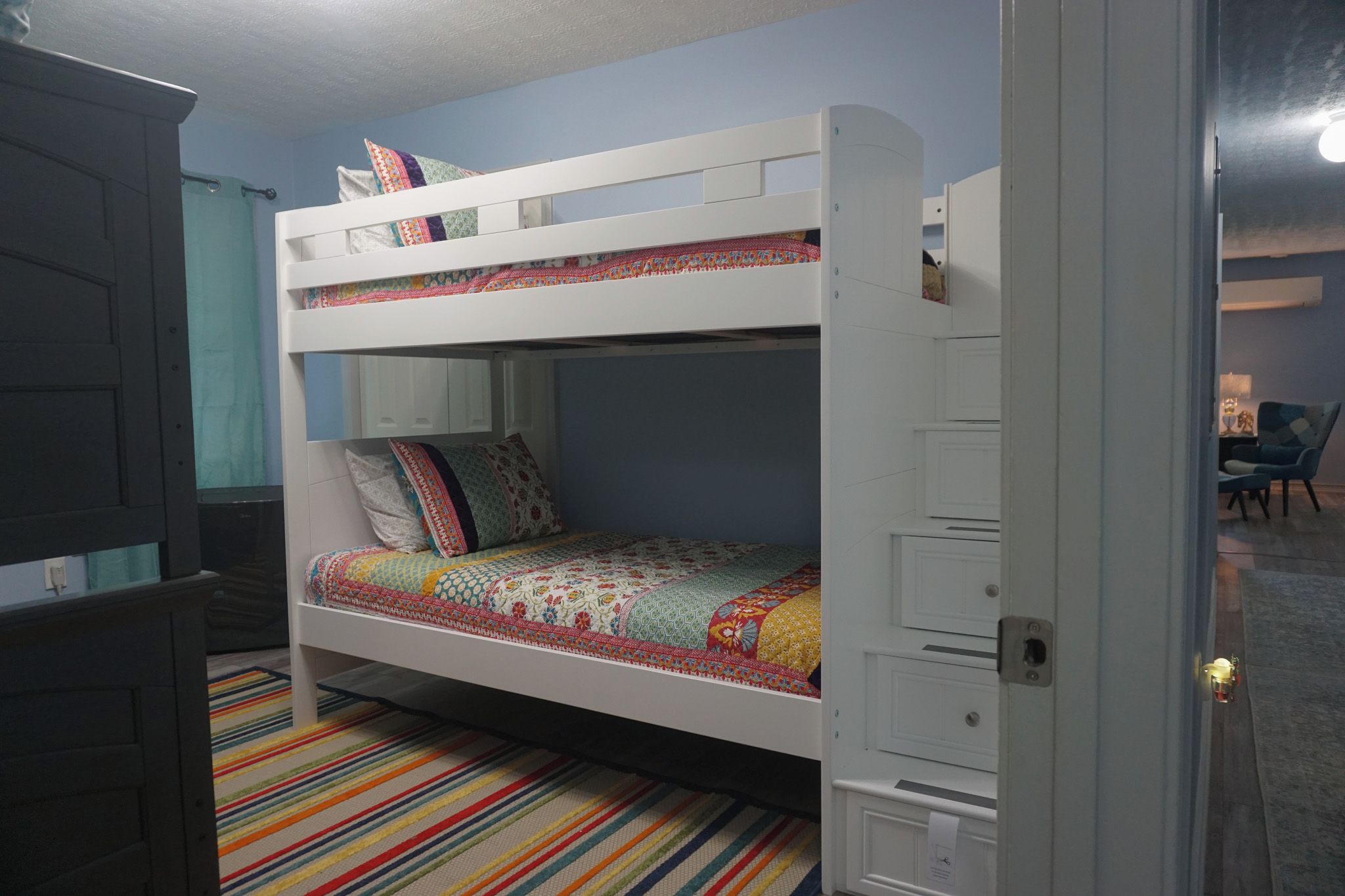 A bunk bed with a colorful quilt, a striped rug, and a door to another room.