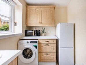 A compact kitchen with wooden cabinets, a washing machine, a microwave, a coffee maker, and a refrigerator. - Charming 2BR Flat Near Holyrood Park & Old Town (Edinburgh)