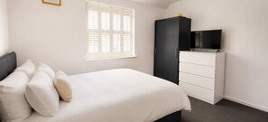 Ein Schlafzimmer mit einem Bett, einem Kleiderschrank, einem Fernseher auf dem Kleiderschrank und einem Fenster mit Jalousien.