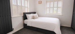 A bedroom with a bed, two windows with shutters, a radiator, and a picture on the wall. - 4BR Home Near Windsor Castle & Legoland (Windsor)