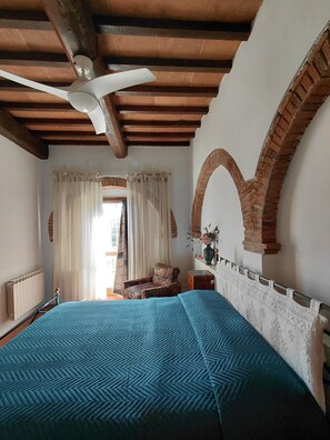 Een slaapkamer met een bed, een plafondventilator, een stoel en een raam met gordijnen.