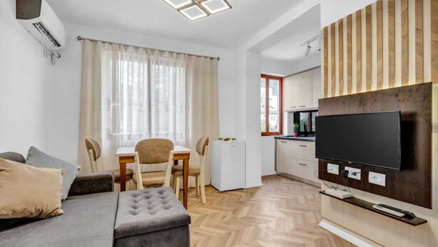 A modern living room with a sofa, ottoman, and a dining area with chairs and a table. A flat-screen TV is mounted on the wall.
