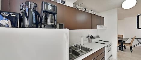 Una cocina moderna con un refrigerador blanco, un lavamanos de acero inoxidable y una estufa. Hay armarios sobre la encimera y una cafetera a la izquierda.
