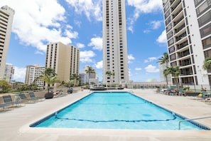 A swimming pool with lounge chairs and palm trees, surrounded by tall residential buildings. - Waikiki Banyan Corner Unit w/ In-Unit W/D & Parking (Honolulu)
