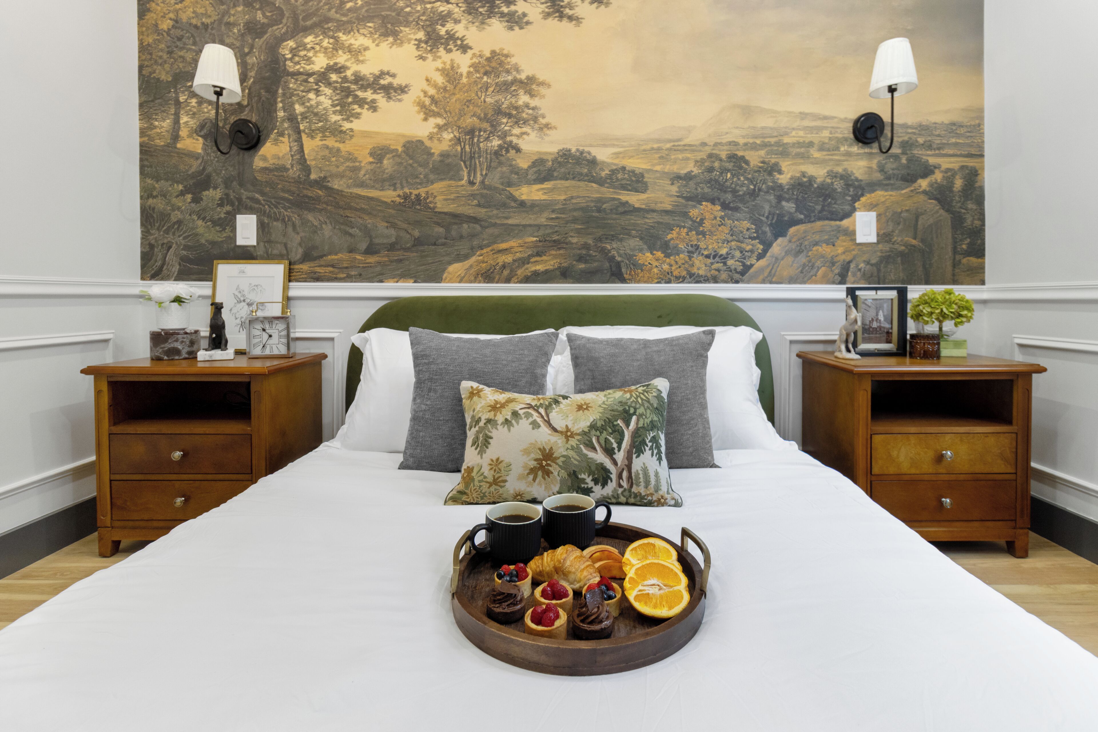Uma cama bem arrumada com uma bandeja de itens de café da manhã, duas mesas de cabeceira com abajures e um quadro grande com paisagem na parede.