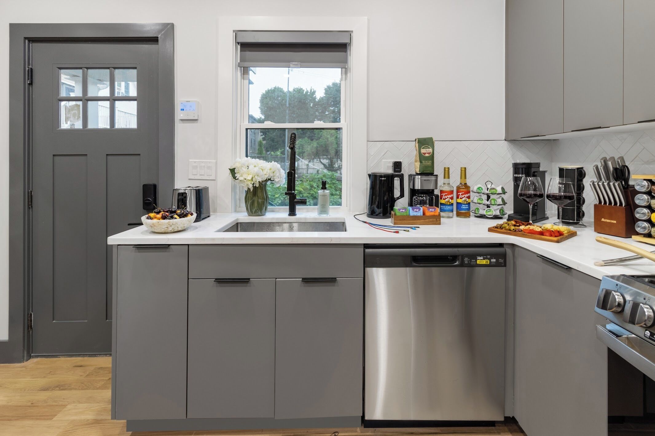 A modern kitchen with grey cabinets, a stainless steel dishwasher, a window with a view, and various kitchen appliances and utensils.