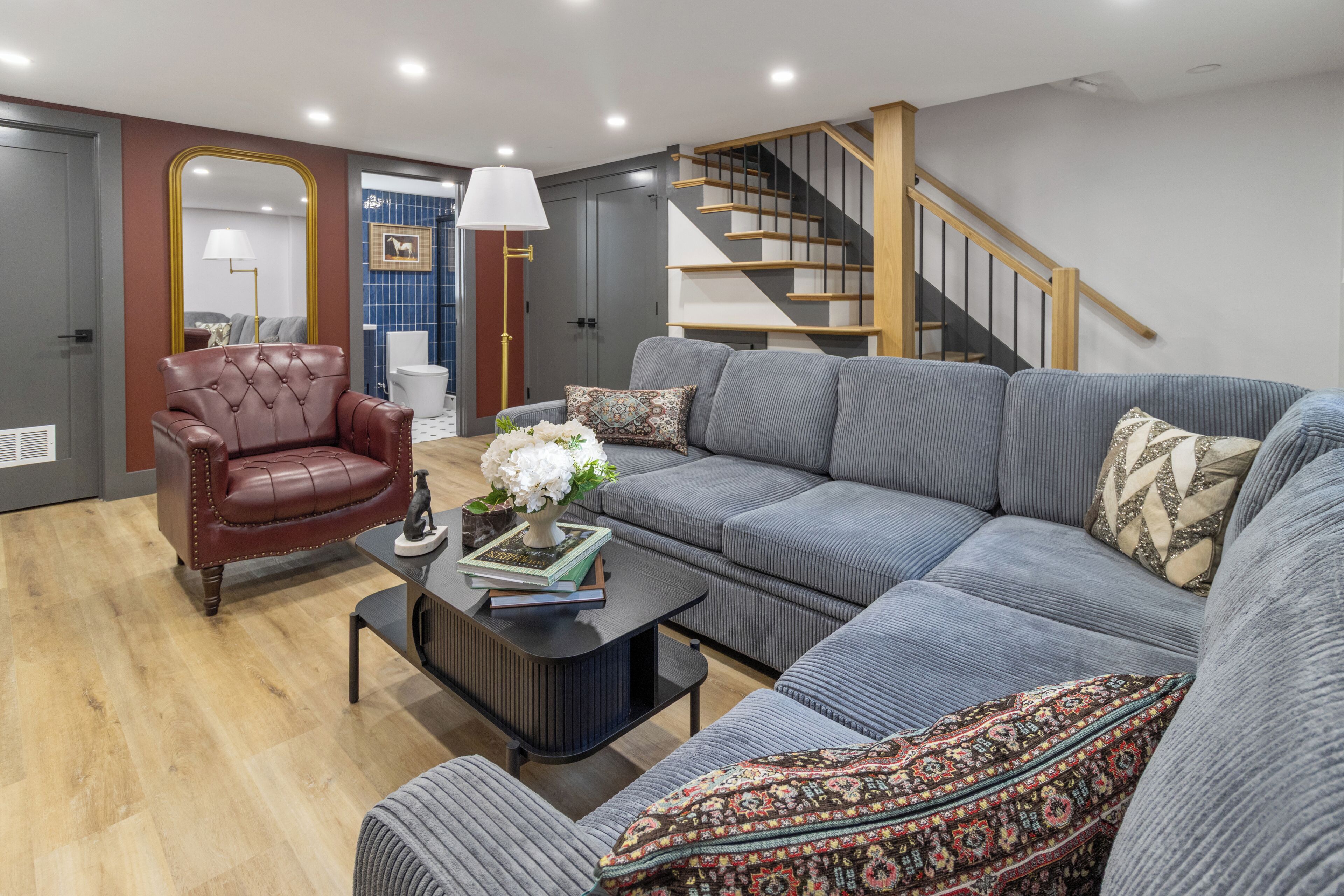 A modern living room with a leather armchair, a grey sofa, a coffee table with books and flowers, and a staircase with wooden railings.