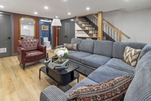 Een moderne woonkamer met een leren fauteuil, een grijze bank, een salontafel met boeken en bloemen, en een trap met houten leuningen.