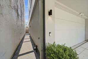 A narrow pathway leading to a garage door, with a small bush in the foreground and a wall-mounted light fixture on the right. - Luxury Houston home located in Houston prime location (Houston)