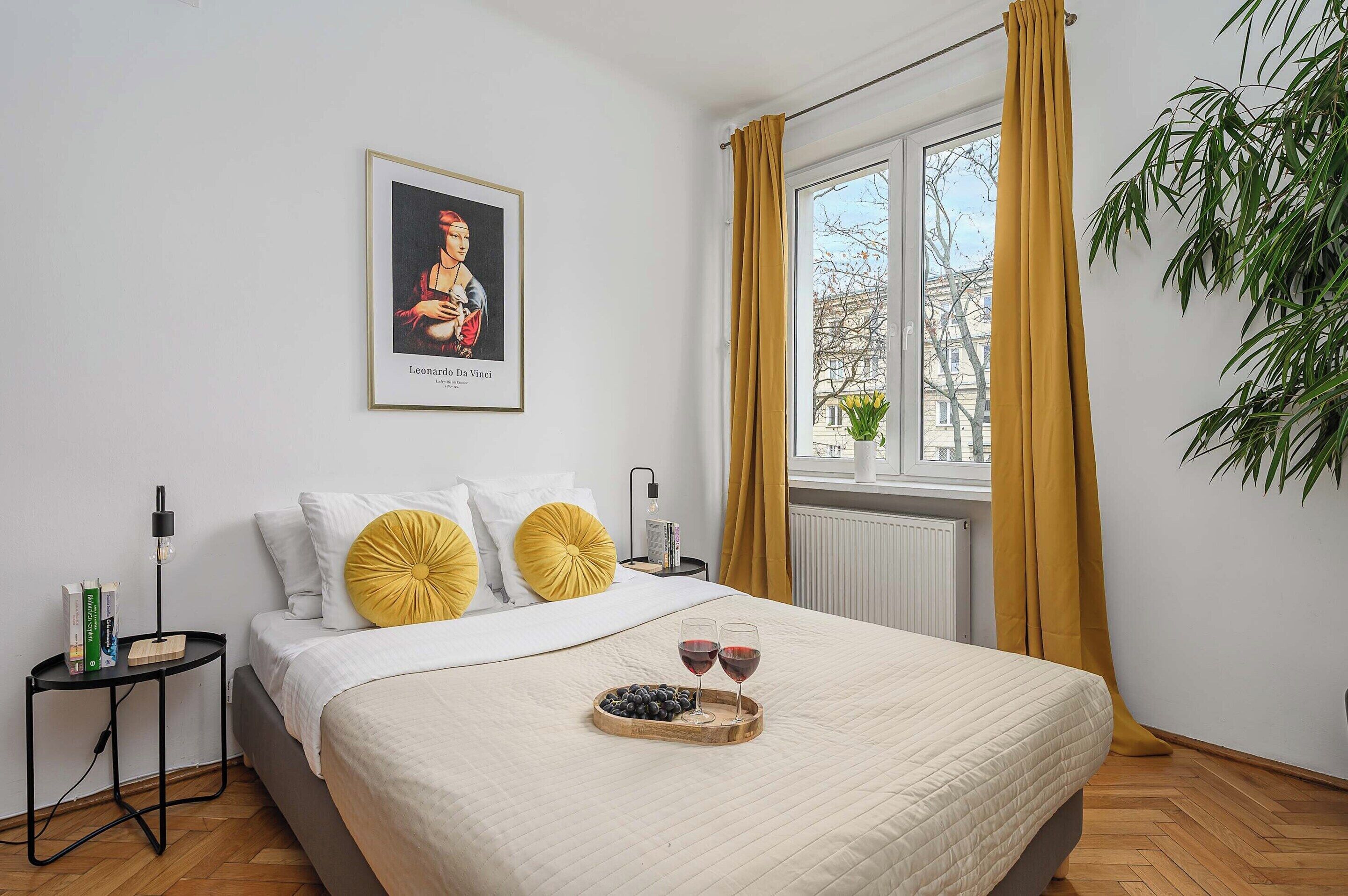 A hotel room with a bed, a bedside table with a lamp, a window with curtains, a plant, and a framed artwork on the wall.