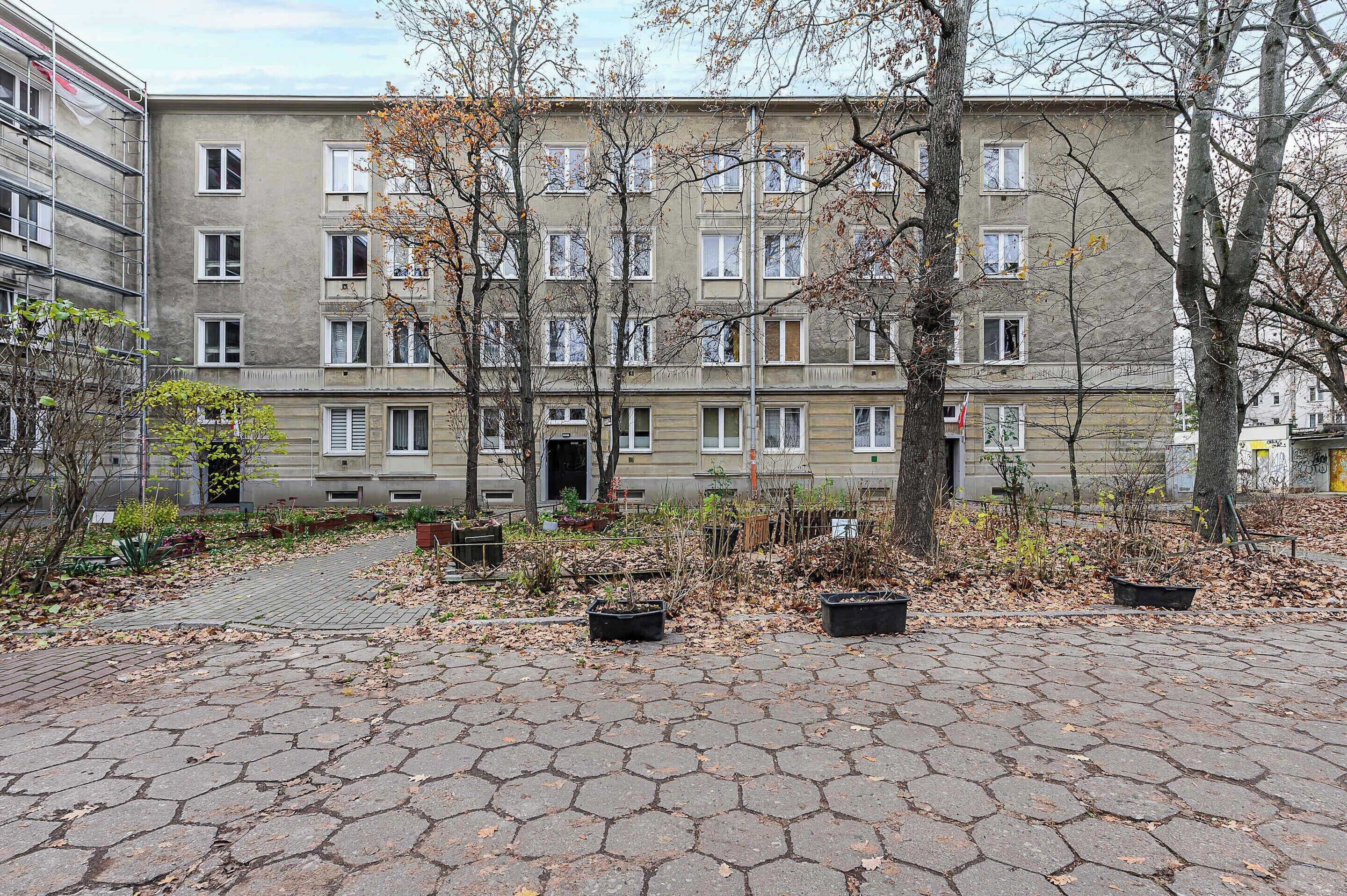A multi-story residential building with a paved courtyard, trees, and a mix of potted plants.