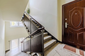 Eine Treppe mit Metallgeländern führt zu einer Zwischenebene, einer Holztür mit Schlüsselloch und einem Fenster mit Blick auf Bäume.