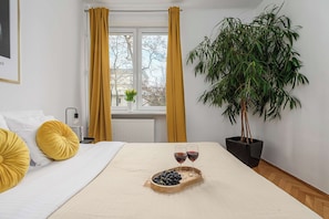 Ein Schlafzimmer mit Bett, Nachttisch mit Lampe, Fenster mit Vorhängen, einem Topfpflanzen und einem Tablett mit Trauben und Weingläsern.