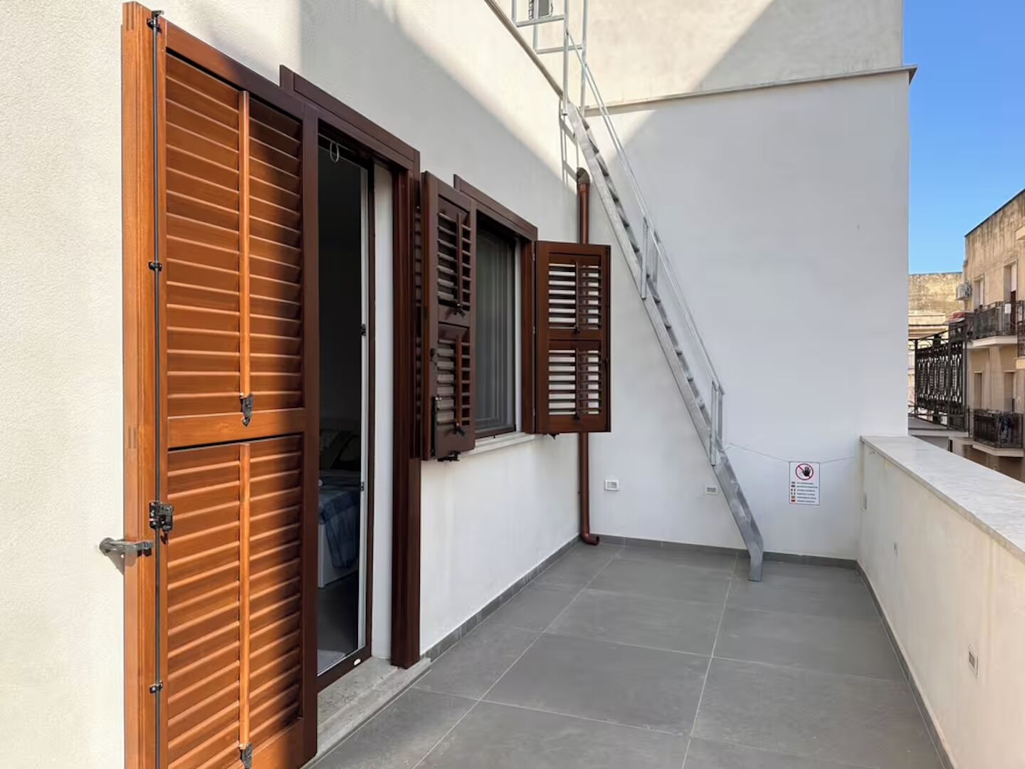 Un balcon avec une porte à volets en bois, une échelle en métal et un sol carrelé.