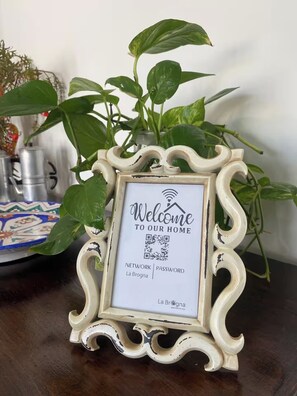 A framed welcome sign with a QR code, placed on a table with a potted plant and a decorative plate. - "La Brogna" Apartment - Downtown Mazara del Vallo (Mazara del Vallo)