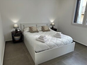 A white bed with a quilted mattress, two nightstands with lamps, and a window with a view of a building. - "La Brogna" Apartment - Downtown Mazara del Vallo (Mazara del Vallo)