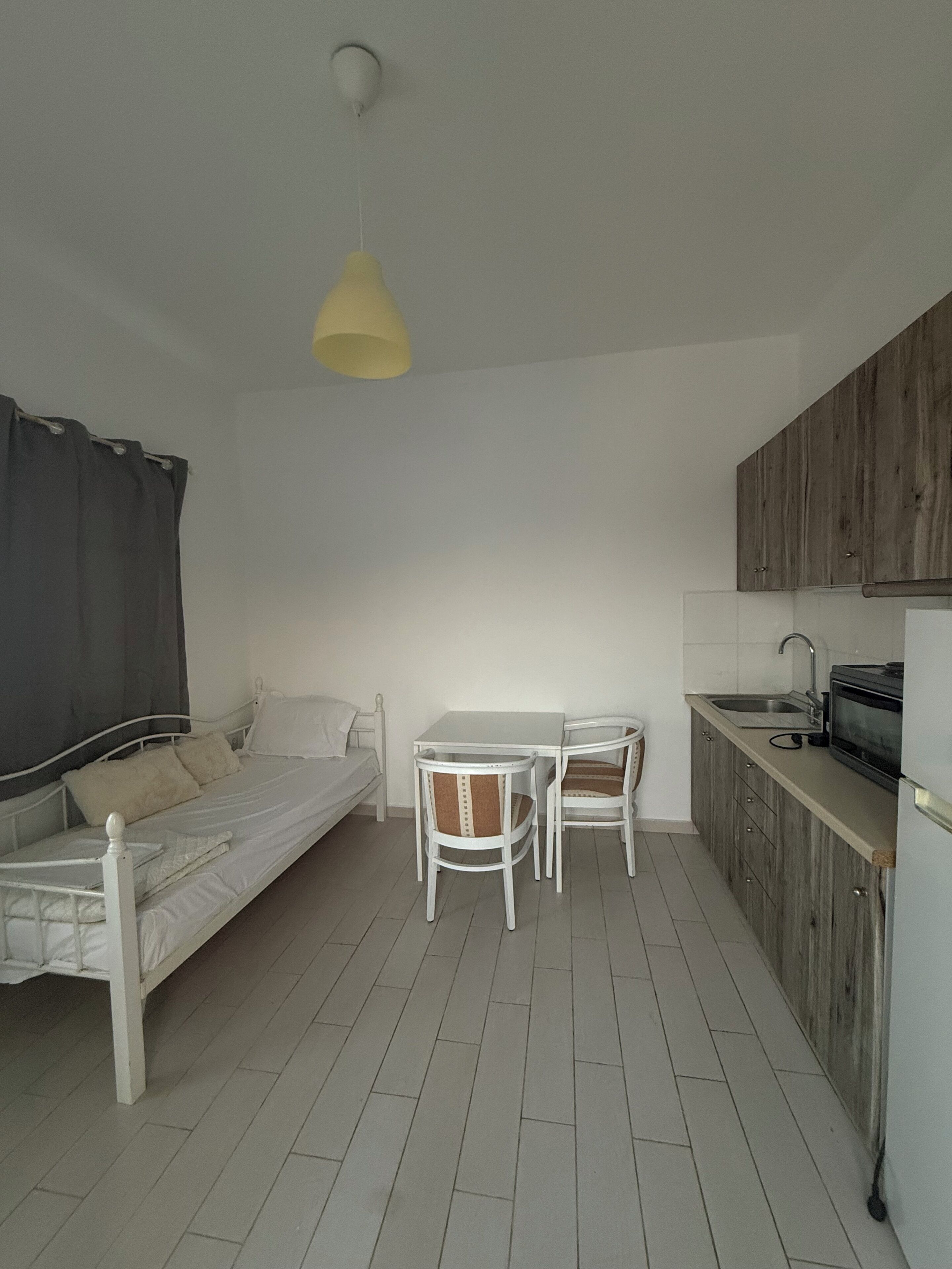 Une pièce de taille modeste comprenant un lit, une petite table à manger avec des chaises, un coin cuisine et un luminaire suspendu.