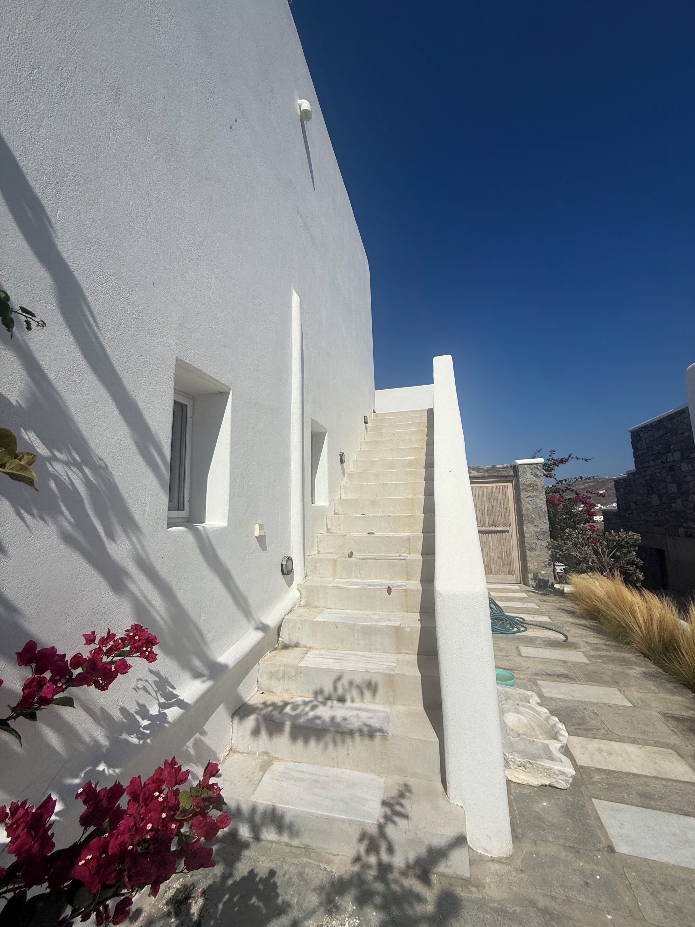 A white building with a staircase leading to a door, surrounded by stone steps and red flowers.