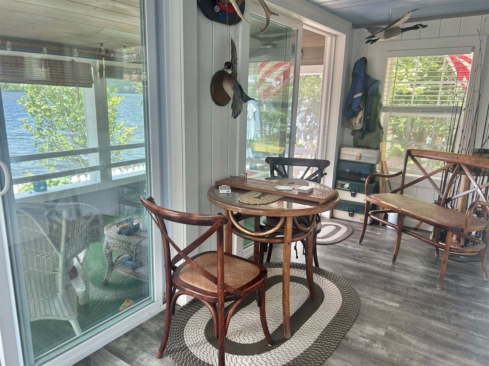 A room with a round wooden table, two chairs, a wicker chair, and a view of the outdoors through large windows.