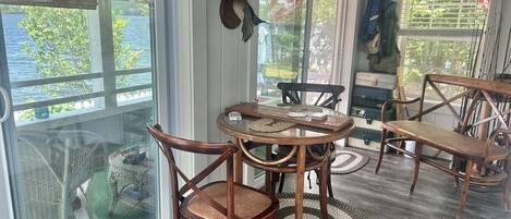 A room with a round wooden table, two chairs, a wicker chair, and a view of the outdoors through large windows.