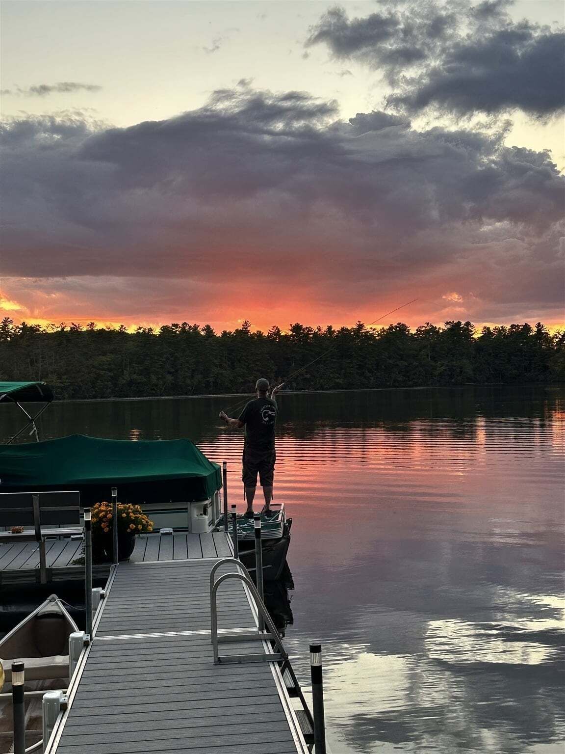 Eine Pier mit einer Person am Rand, die einen ruhigen See mit bewaldeter Uferlinie und einem Sonnenuntergang im Hintergrund betrachtet.