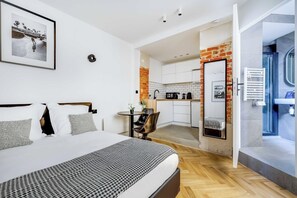 Habitación de hotel moderna con una cama grande, una pequeña mesa redonda y una cocina visible a través de una puerta abierta.