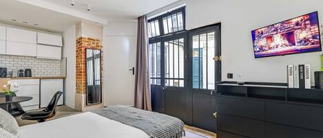 A modern hotel room with a bed, a small kitchen area, a dining table, and a large window.