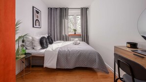 A bedroom with a bed, a desk, a chair, a radiator, a window with curtains, and a plant. - Wola Flat Near City Center (Warsaw)