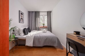 Ein Schlafzimmer mit einem Bett, einem Schreibtisch, einem Stuhl, einem Heizkörper, einem Fenster mit Vorhängen und einer Pflanze.