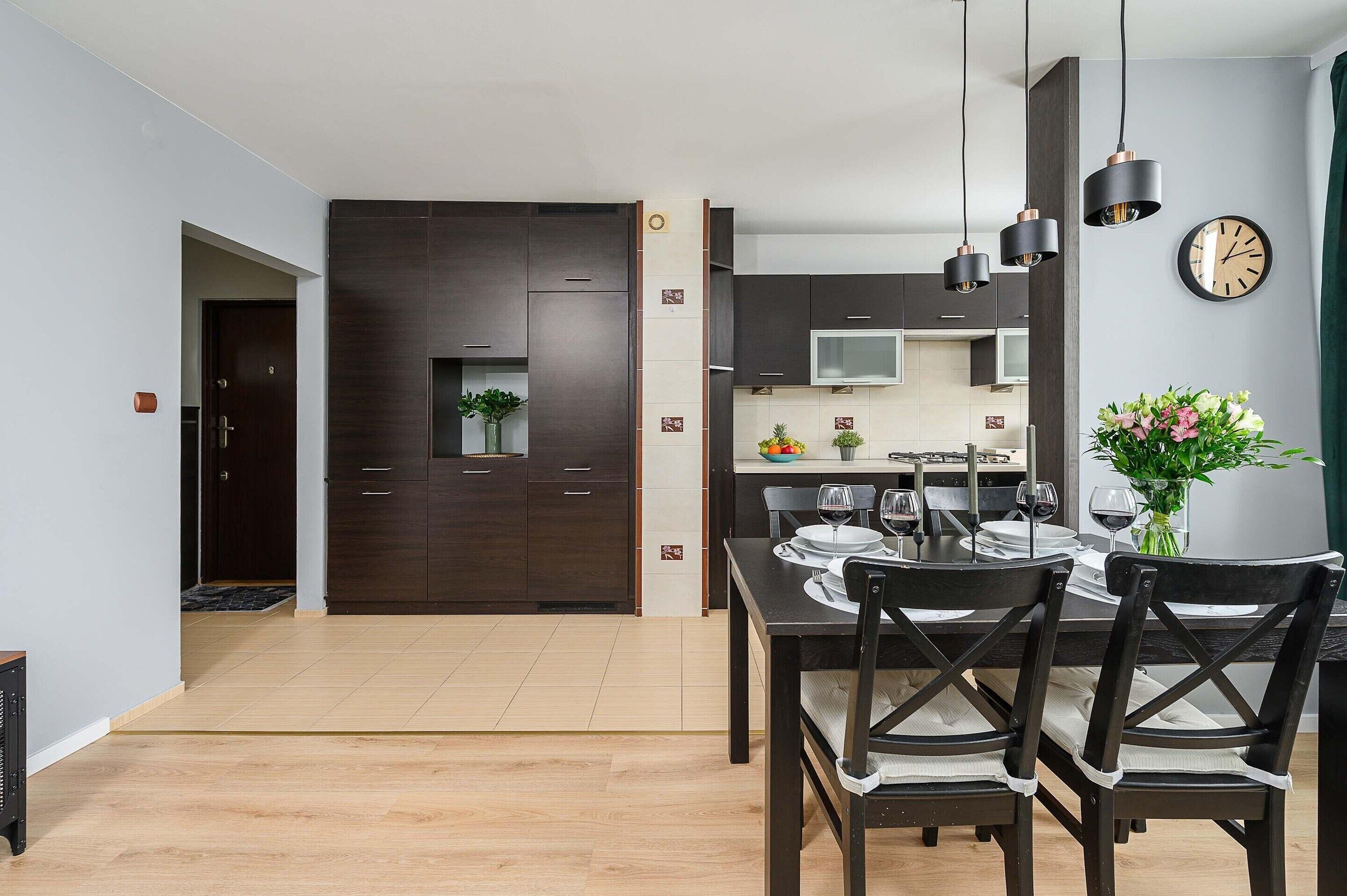 A modern kitchen with dark cabinetry, a dining table set for four, and a clock on the wall.