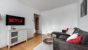 Uma sala de estar moderna com uma televisão de tela plana exibindo 'NETFLIX', um sofá cinza com almofadas vermelhas e brancas, uma mesa de centro de vidro com revistas e um armário branco com gavetas.