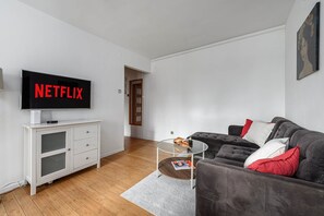 Uma sala de estar moderna com uma televisão de tela plana exibindo 'NETFLIX', um sofá cinza com almofadas vermelhas e brancas, uma mesa de centro de vidro com revistas e um armário branco com gavetas.