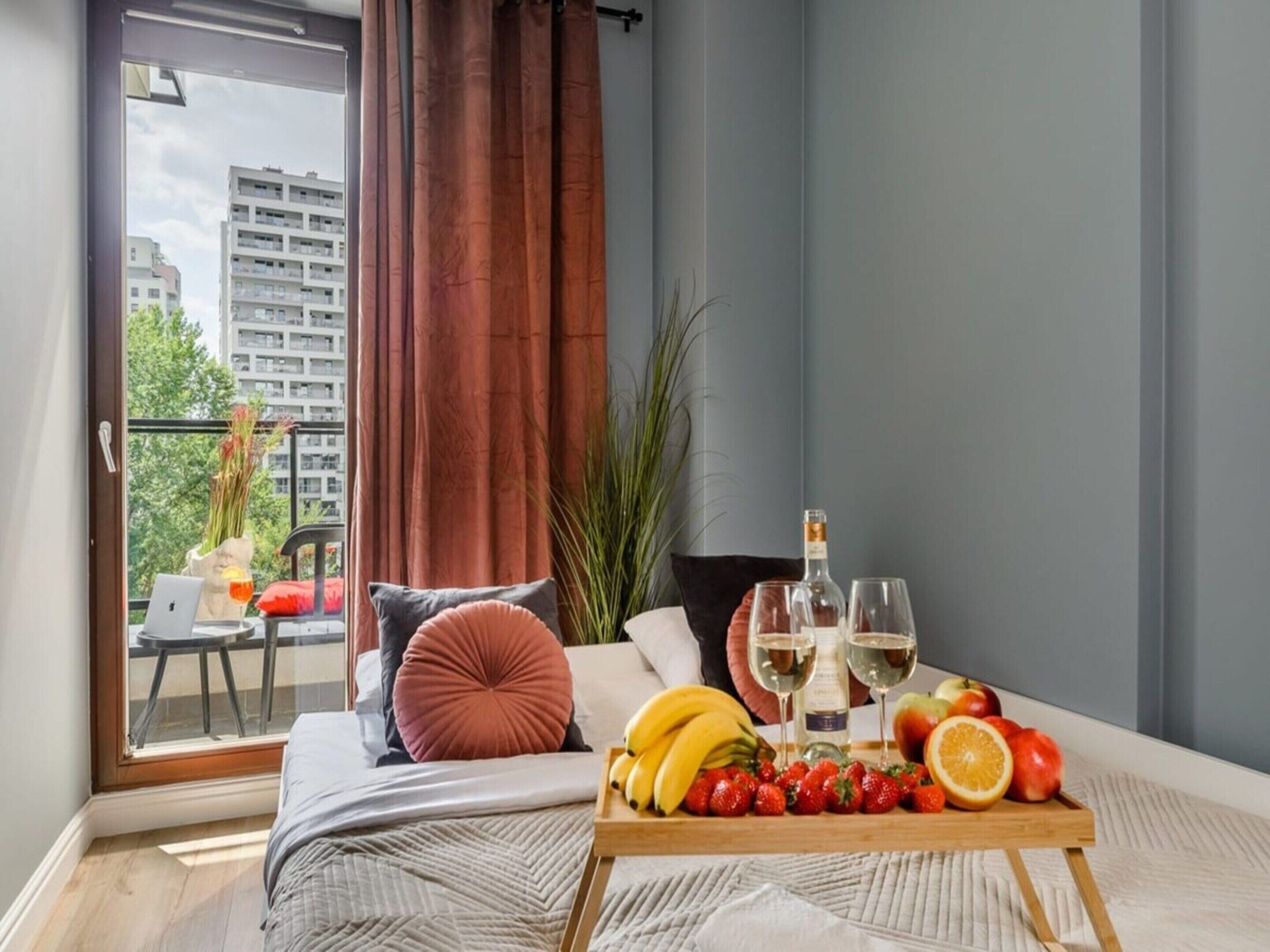 Une chambre à coucher moderne avec un lit, une petite table sur laquelle sont disposés des fruits et une bouteille de vin, et une vue sur les bâtiments par la fenêtre.
