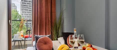 A modern bedroom with a bed, a small table with fruit and wine, and a view of buildings through a window.