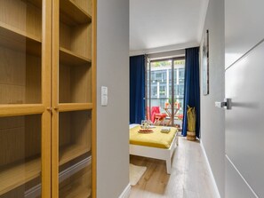 Une pièce comprenant un lit, une table recouverte d’une housse de couette jaune, une fenêtre avec des rideaux bleus et une armoire en bois.