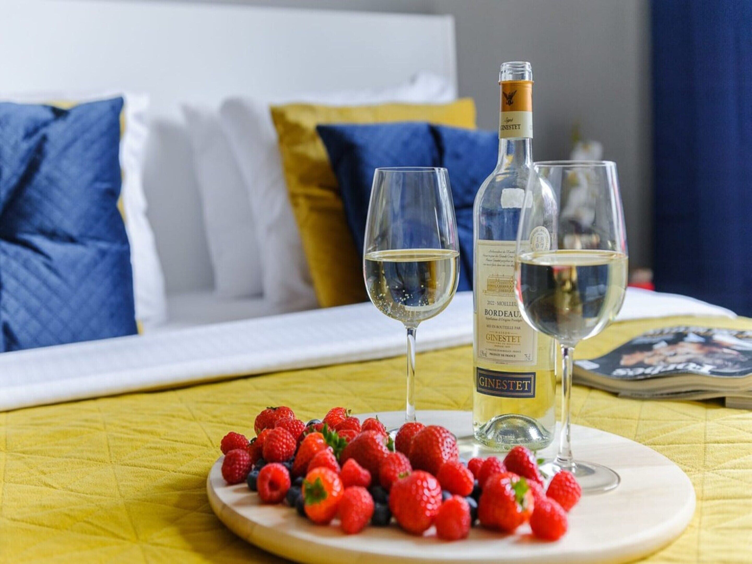 Una cama con ropa de cama azul y blanca, una botella de vino, dos copas y un plato de fresas sobre un mantel amarillo.