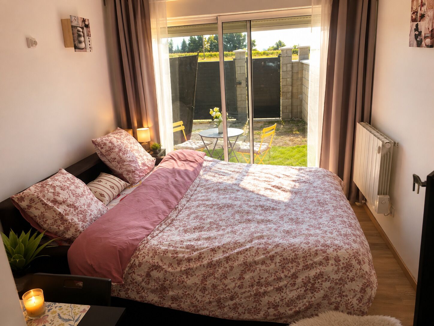 A bedroom with a bed, a bedside table with a lamp, a small table with a candle, and a view of a garden through a sliding door.