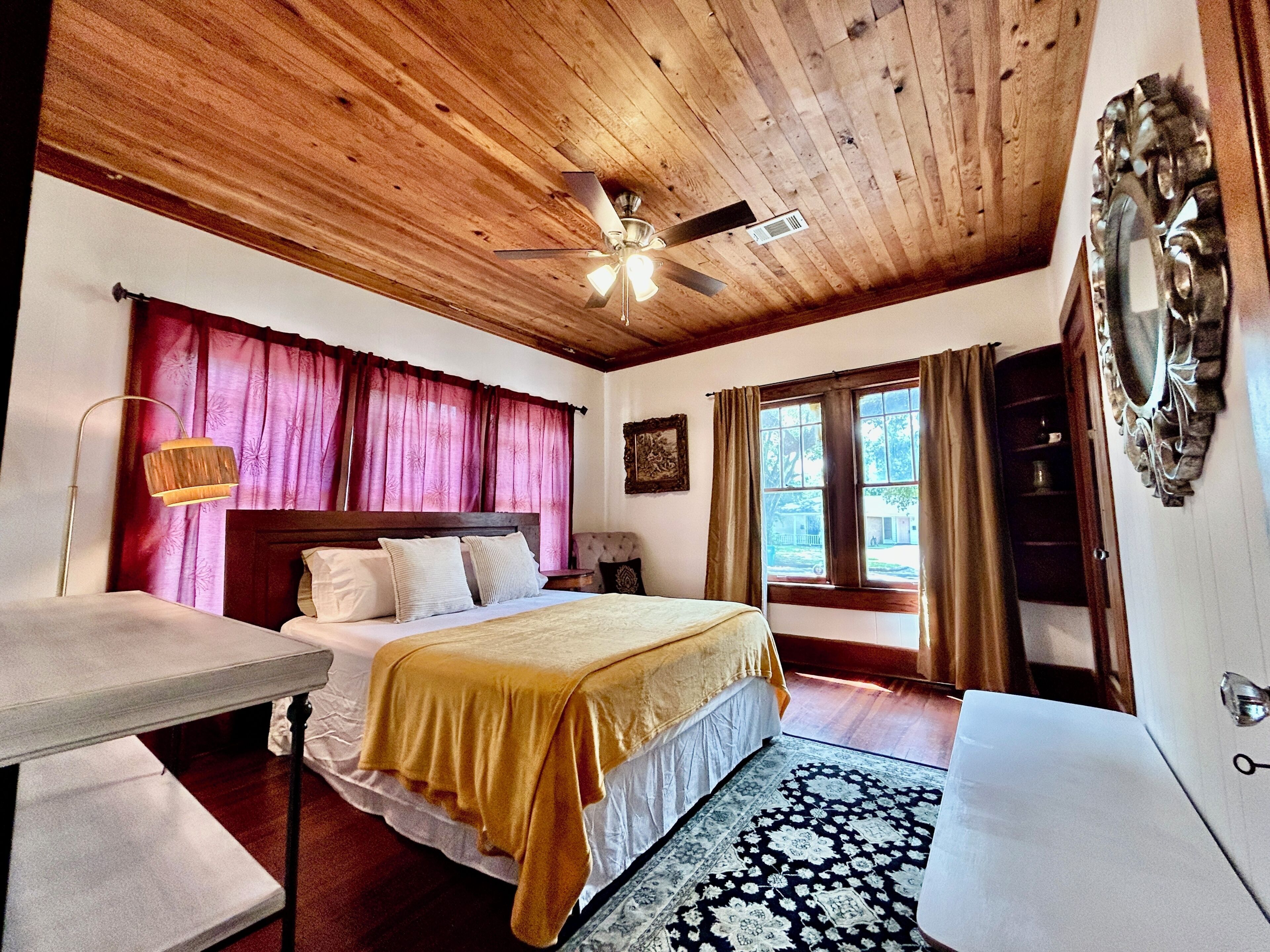 Quarto com cama, escrivaninha, ventoinha no teto e janela com cortinas.