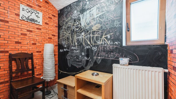 Ein Zimmer mit Backsteinwand, einem Holzstuhl, einem Heizkörper und einer Tafel mit Schrift.