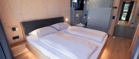 Une chambre moderne avec un plancher en bois, un grand lit recouvert de draps blancs, une tête de lit sombre et une cabine de douche en verre.