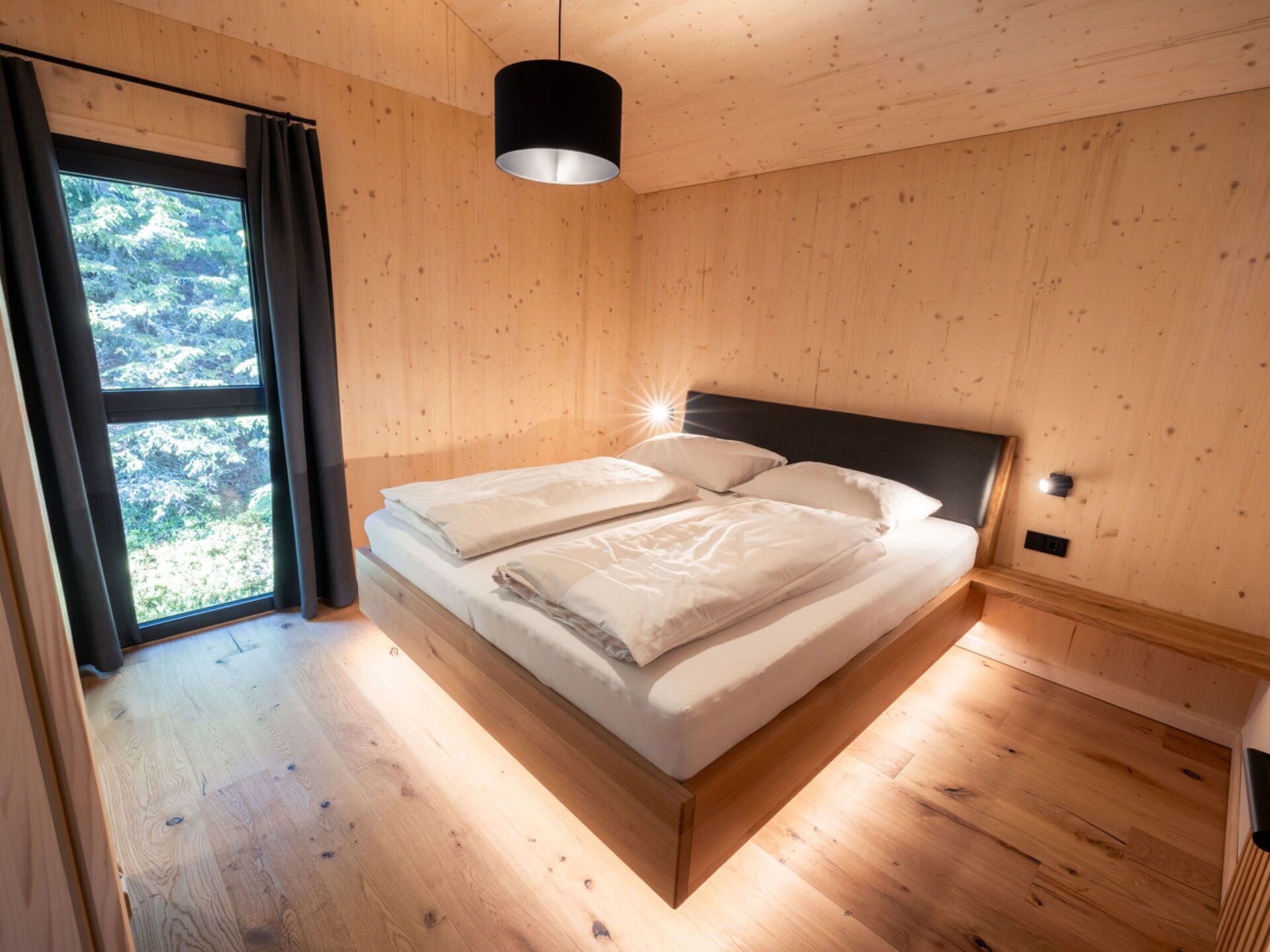 Een slaapkamer met houten vloer, een bed met witte lakens, een zwart hoofdeinde en een raam met gordijnen.