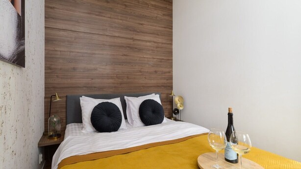 Ein Hotelzimmer mit einem hölzernen Kopfteil, einem Bett mit gelber Tagesdecke, zwei schwarzen Kissen, einem Nachttisch mit Lampe und einem Tablett mit einer Weinflasche und Gläsern.