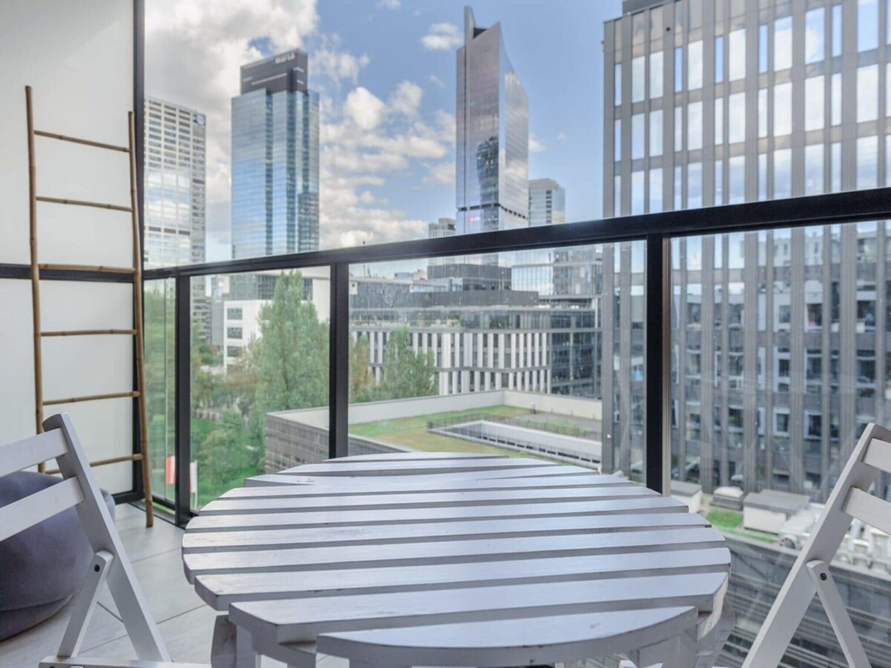 Un balcón moderno con una mesa redonda y sillas, con vista a un paisaje urbano lleno de rascacielos.