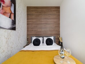 Ein Hotelzimmer mit einem Bett, einem gelben Tisch mit Flasche und Gläsern sowie einem hölzernen Kopfende.