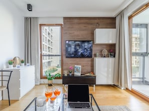 A modern living room with a flat-screen TV, a laptop on a glass table, and a view of a building outside. - Warsaw Flat With Quiet Loggia (Warsaw)